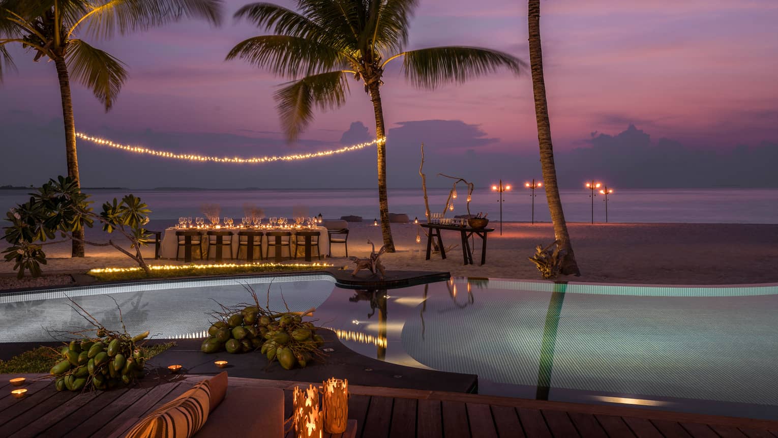 String of lights between palm tree over private dining table on beach by swimming pool at sunset