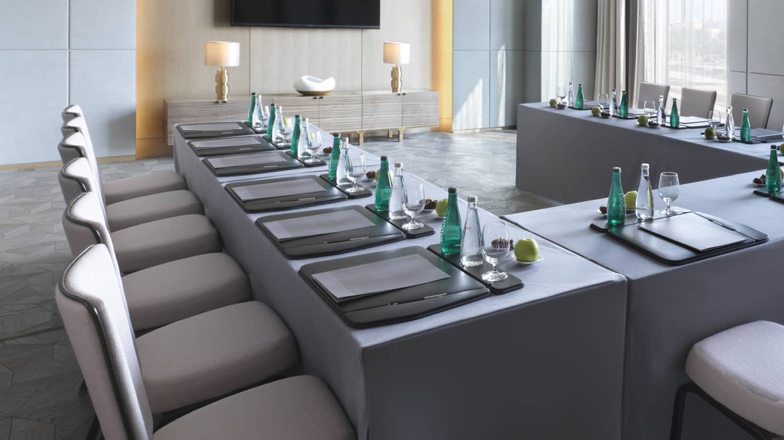 U-shaped desks with water, stationery and fruit in light-filled meeting room facing TV screen