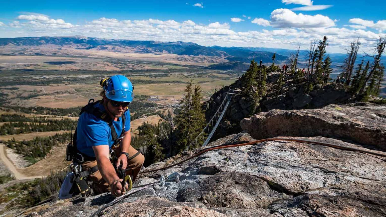 A man in blue clothes and blue helmet rock climbing, an open vista of green land and mountains can be seen behind him.
