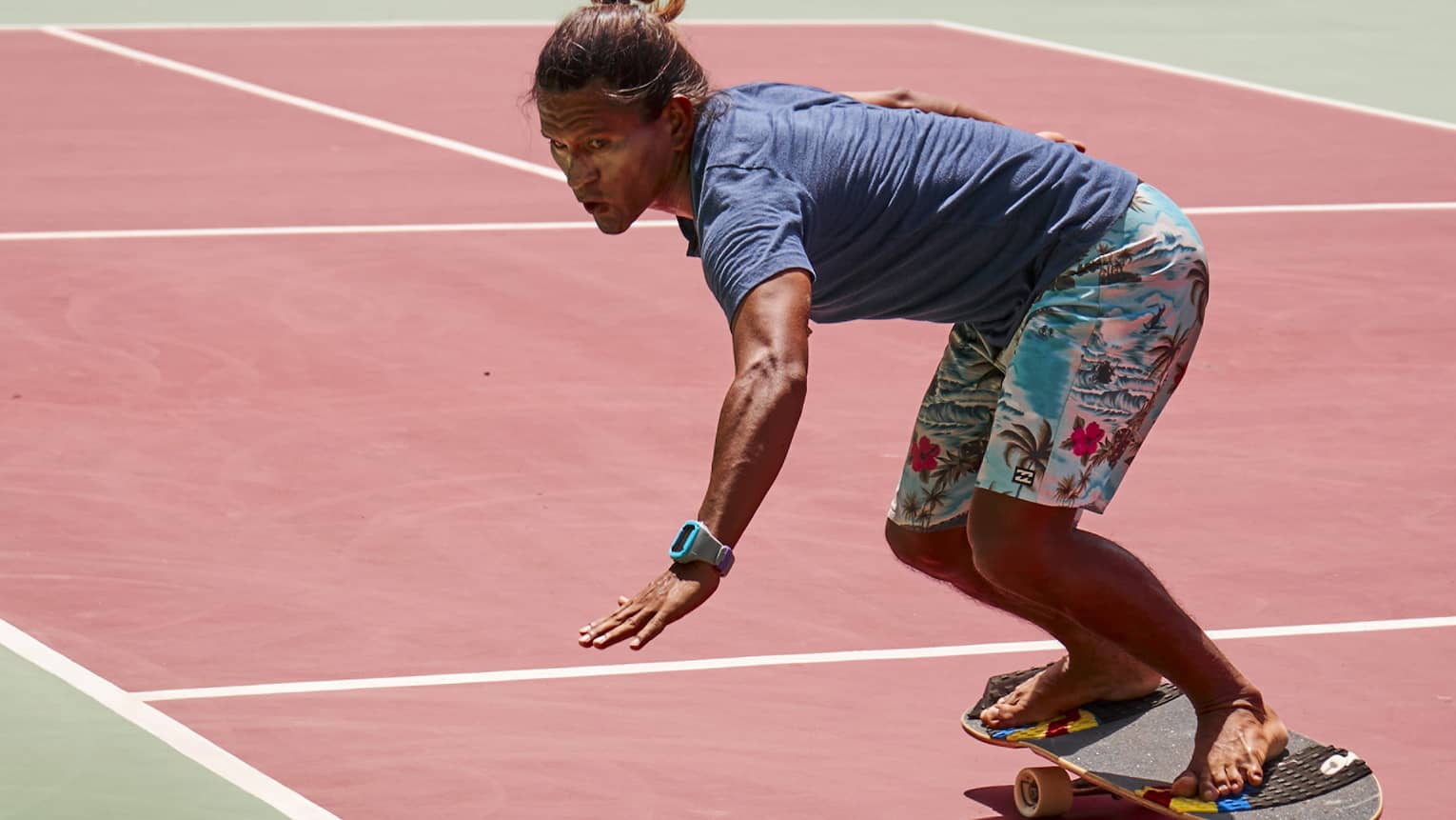 Person wearing light-blue, Hawaiian-print board shorts and a navy t-shirt rides a skateboard around a cone on a tennis court