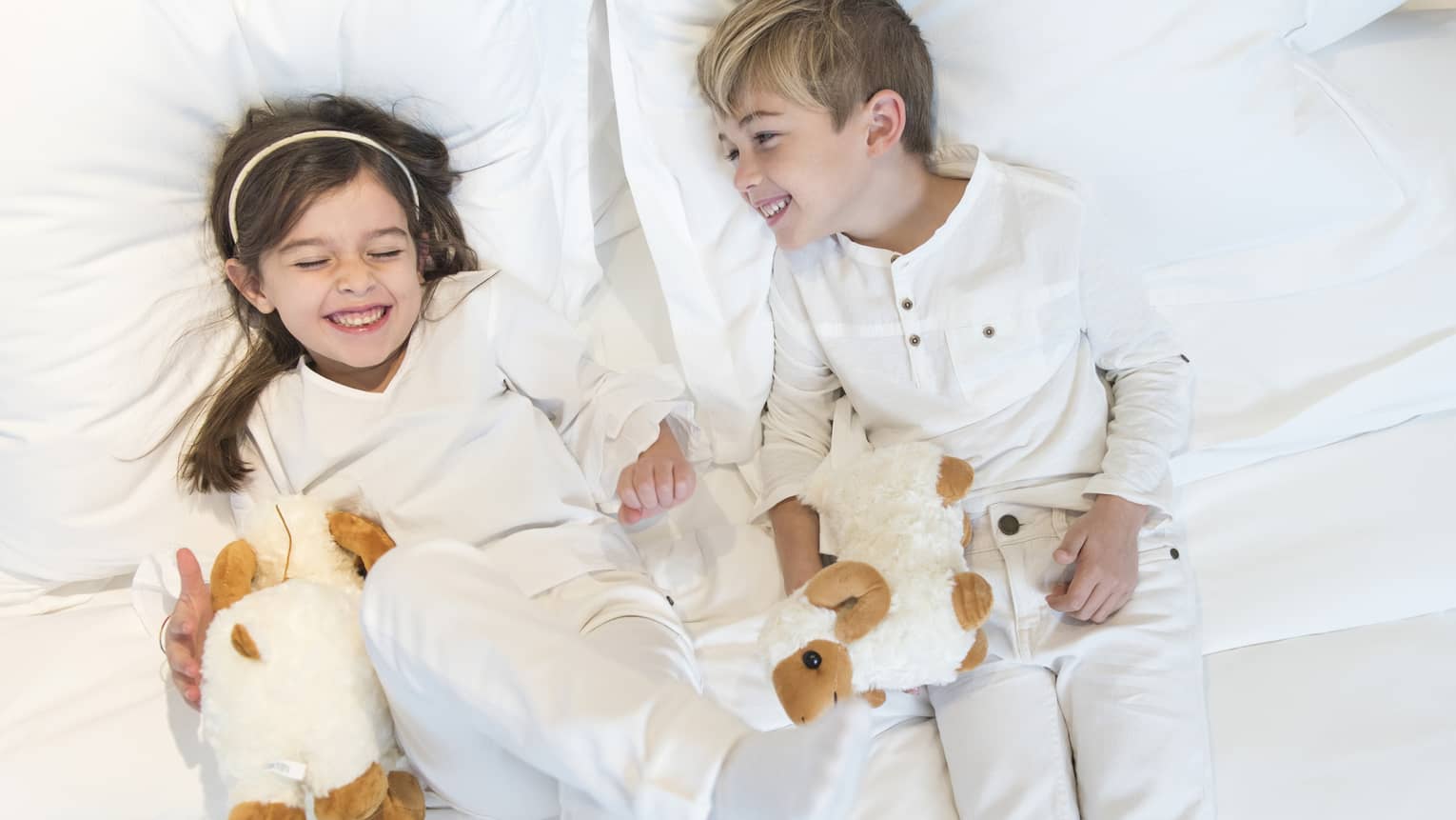 Aerial view of two children wearing white pyjamas with white stuffed animals lying on bed, laughing