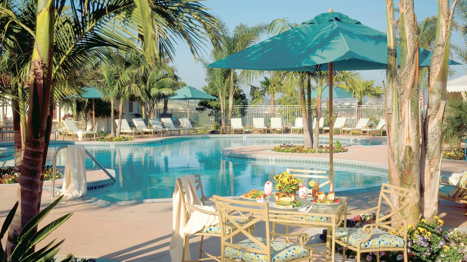 Outdoor pool and pool terrace, white lounge chairs, a square table and four chairs, turquoise umbrella, palms