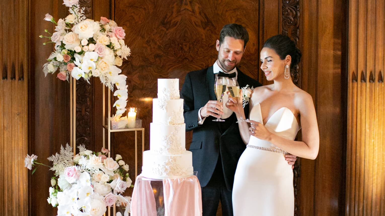 A bride and groom looking at a tall wedding cake surrounded by flowers.