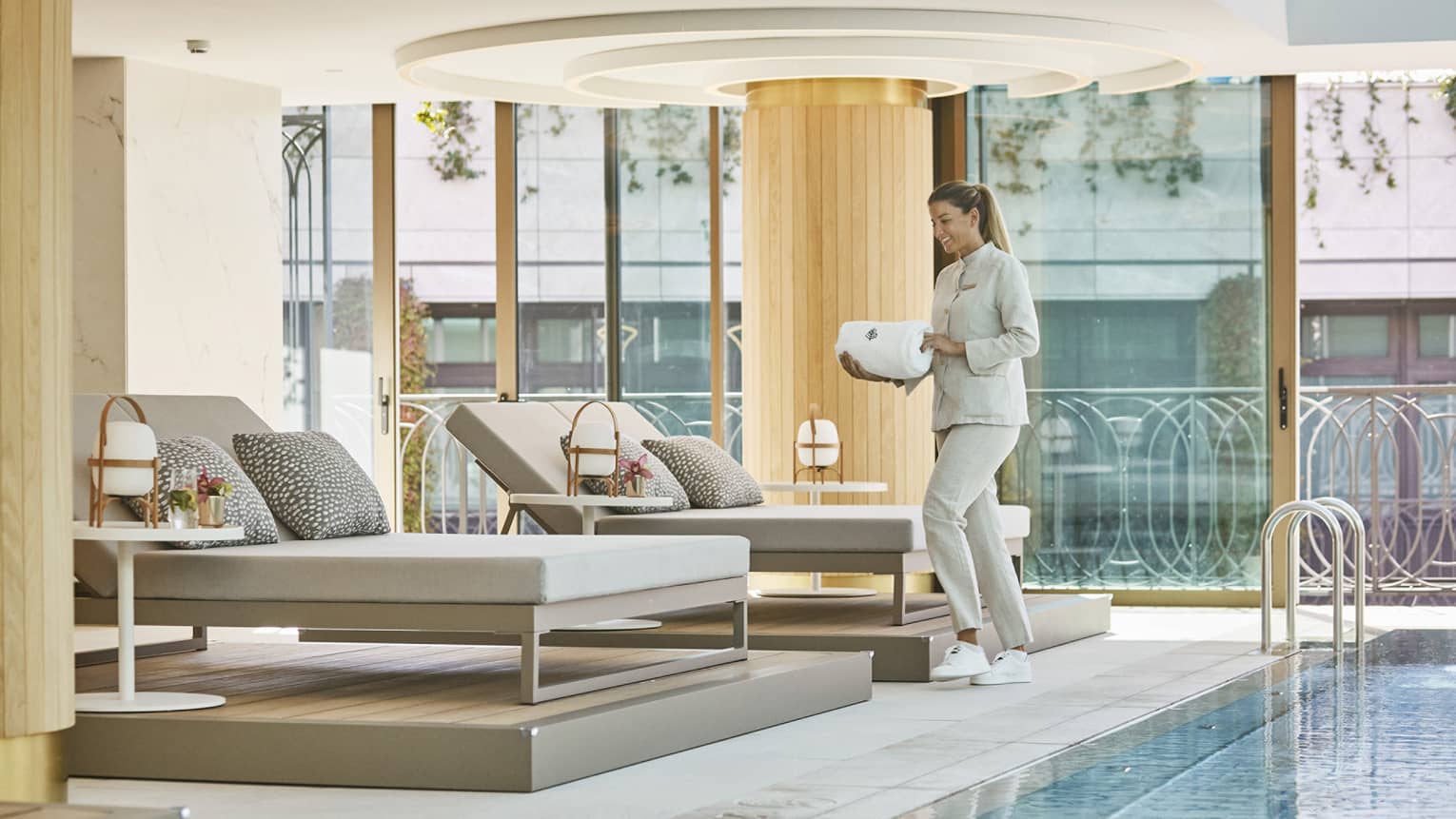 A woman carrying white towels to cushioned lounge seat next to indoor pool