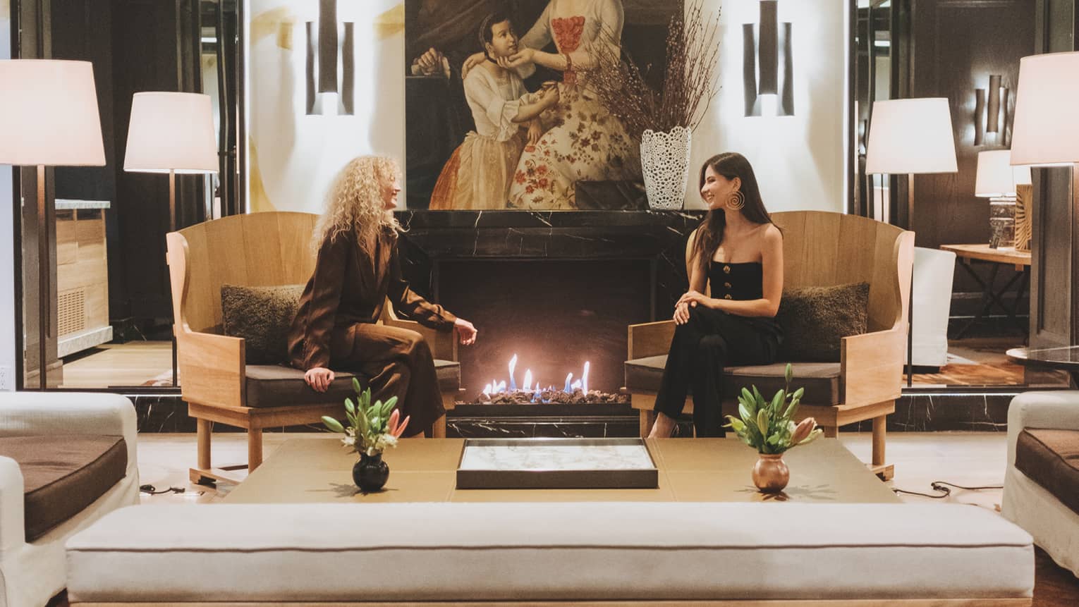 Two women sit in large tan wingback chairs in front of fireplace in chic living room