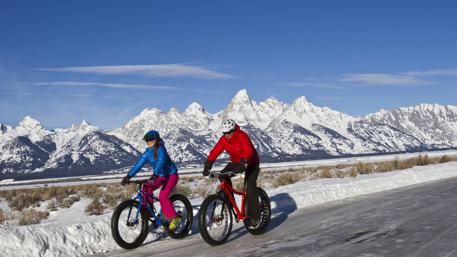 A man and woman bike down a path, snow-capped mountains and blue sky in the background