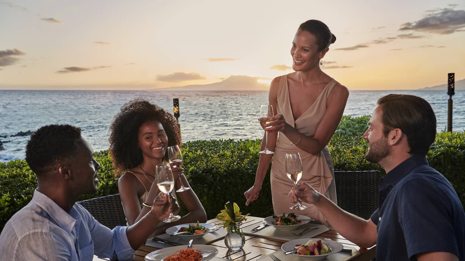 Two couples dine outside at Ferraro’s Bar E Ristorante, overlooking Wailea Beach. The sun is setting as they raise a toast. 