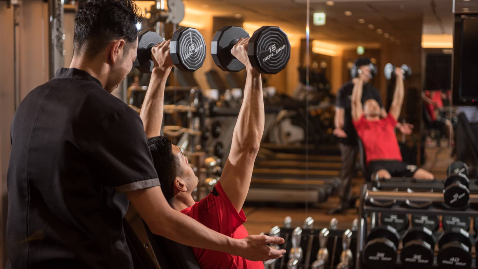 Man in chair in front of mirror lifts two 15 lb. weights above his head while personal trainer assists