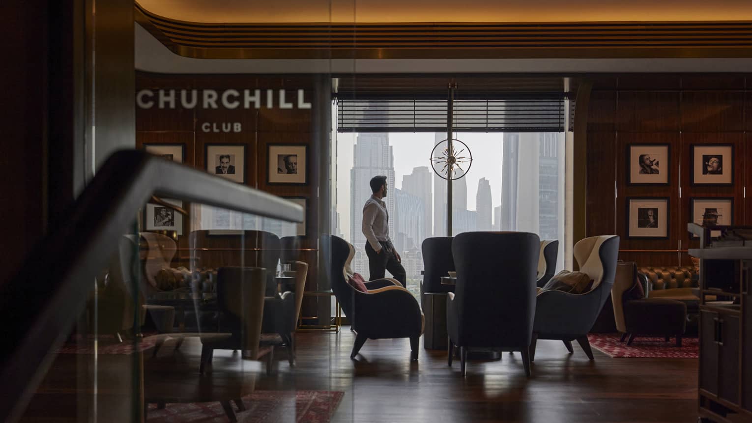 Man stands by a floor-to-ceiling window looking out on the city skyline in a dimly lit cigar bar filled with club chairs and leather sofas