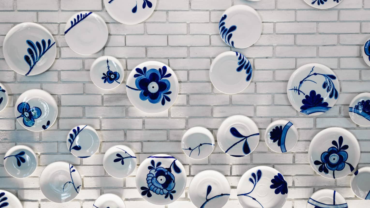 Wall art made up of various white plates painted with blue leaves and flowers hang on a white brick wall
