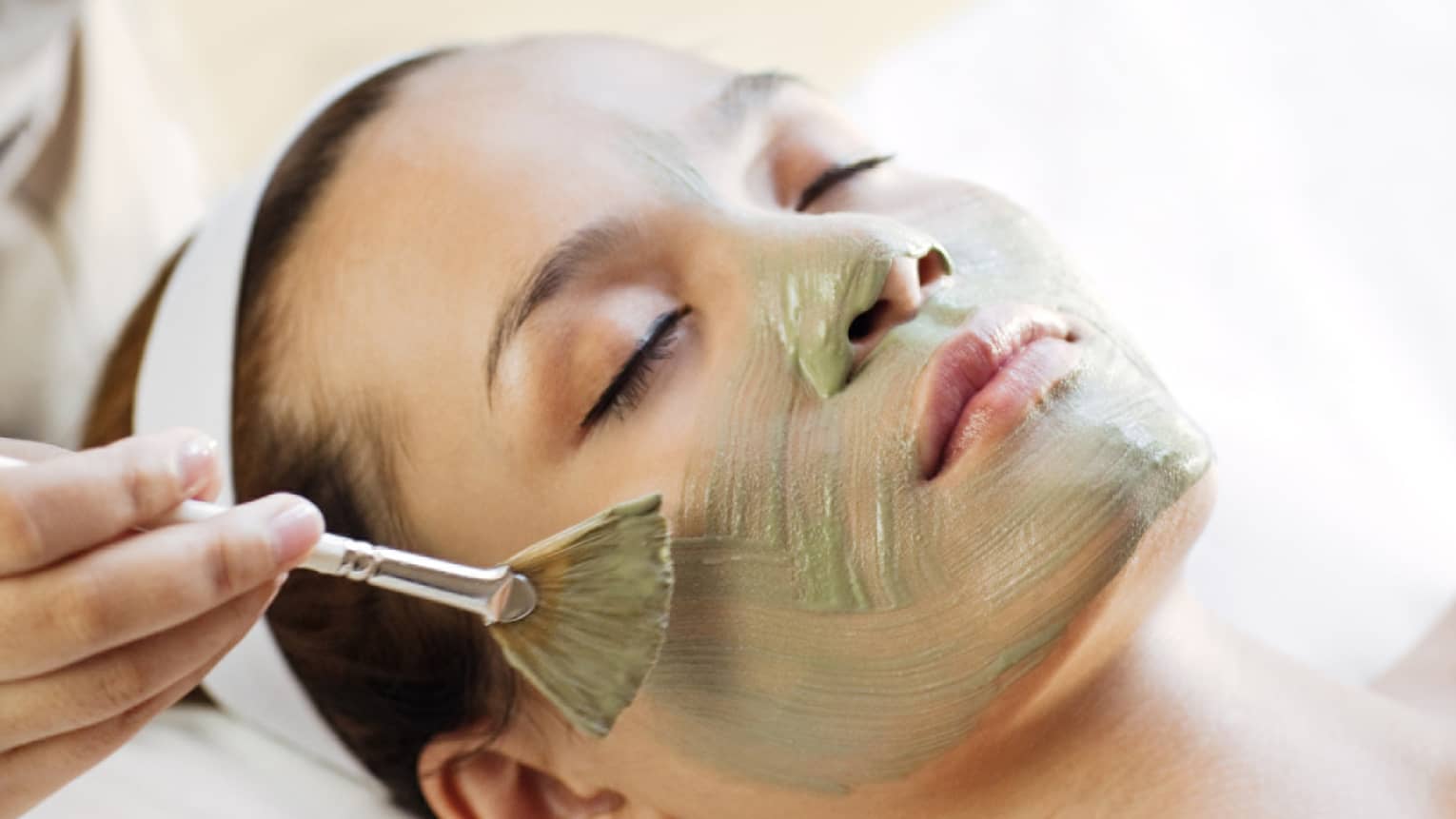 Spa staff holding brush paints green mask on woman's cheeks, chin, nose 