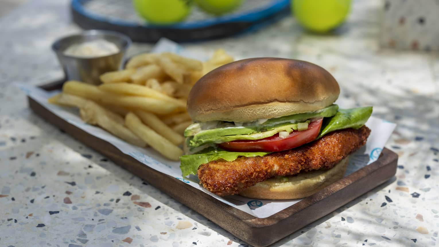 Crispy chicken sandwich with tomato, lettuce, avocado on a bun served with a side of ranch dressing and fries on a rectangular wooden plate