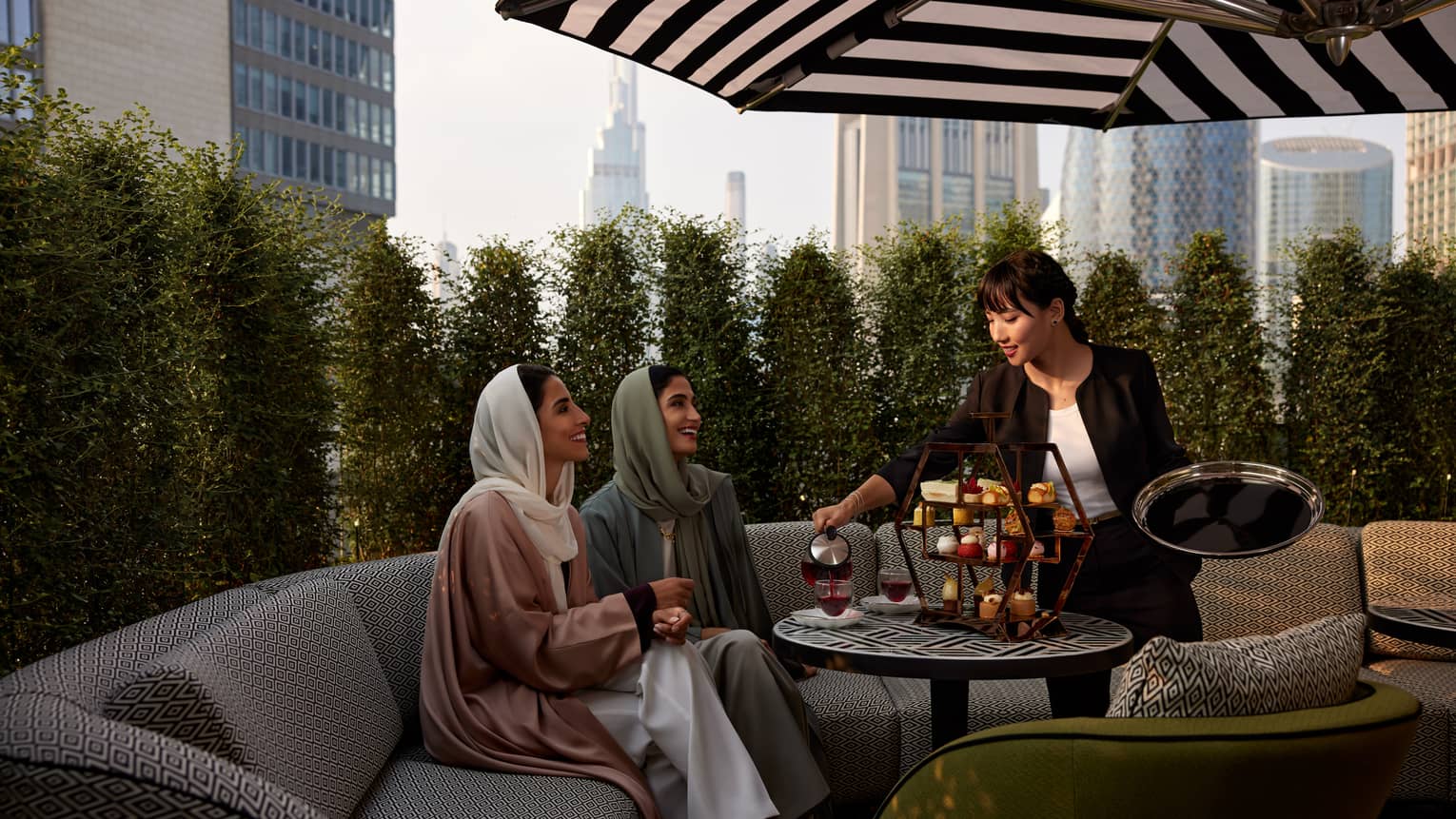 Two women sit on a beige patterned sofa beneath a black-and-white patio umbrella as a waitress serves them tea and pastries