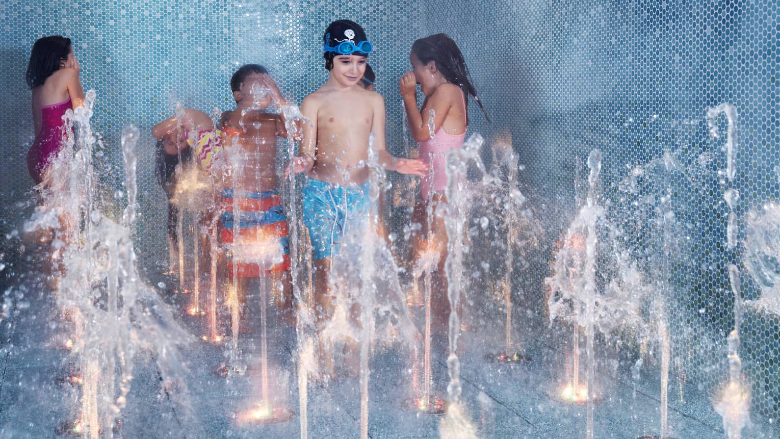 Six children play in a blue-tiled splash pad as water rises up from different holes in the floor