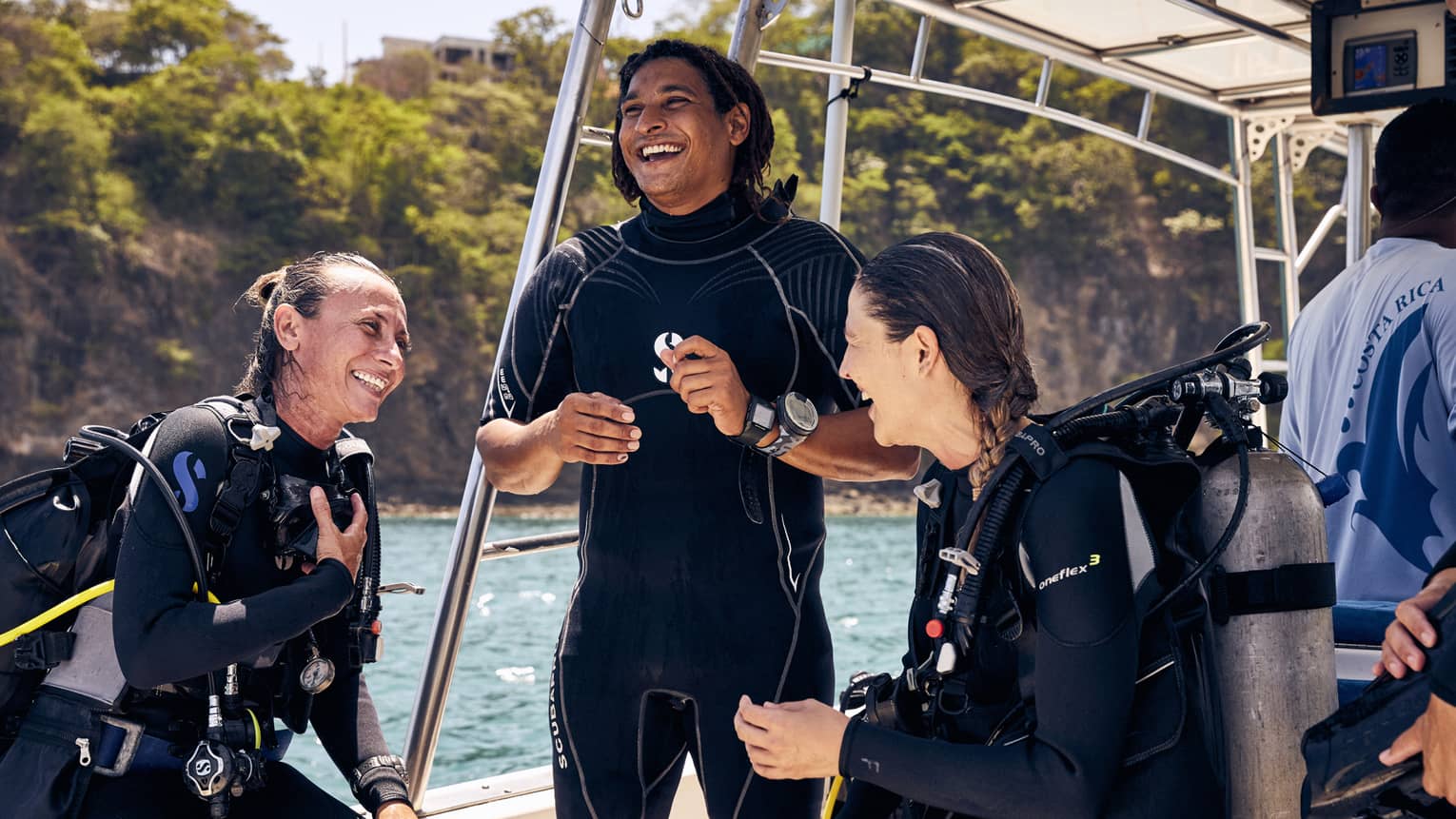 Three scuba-divers in wetsuits laugh under the canopy of a boat on sparkling turquoise water amid a rocky tree-covered cliff.