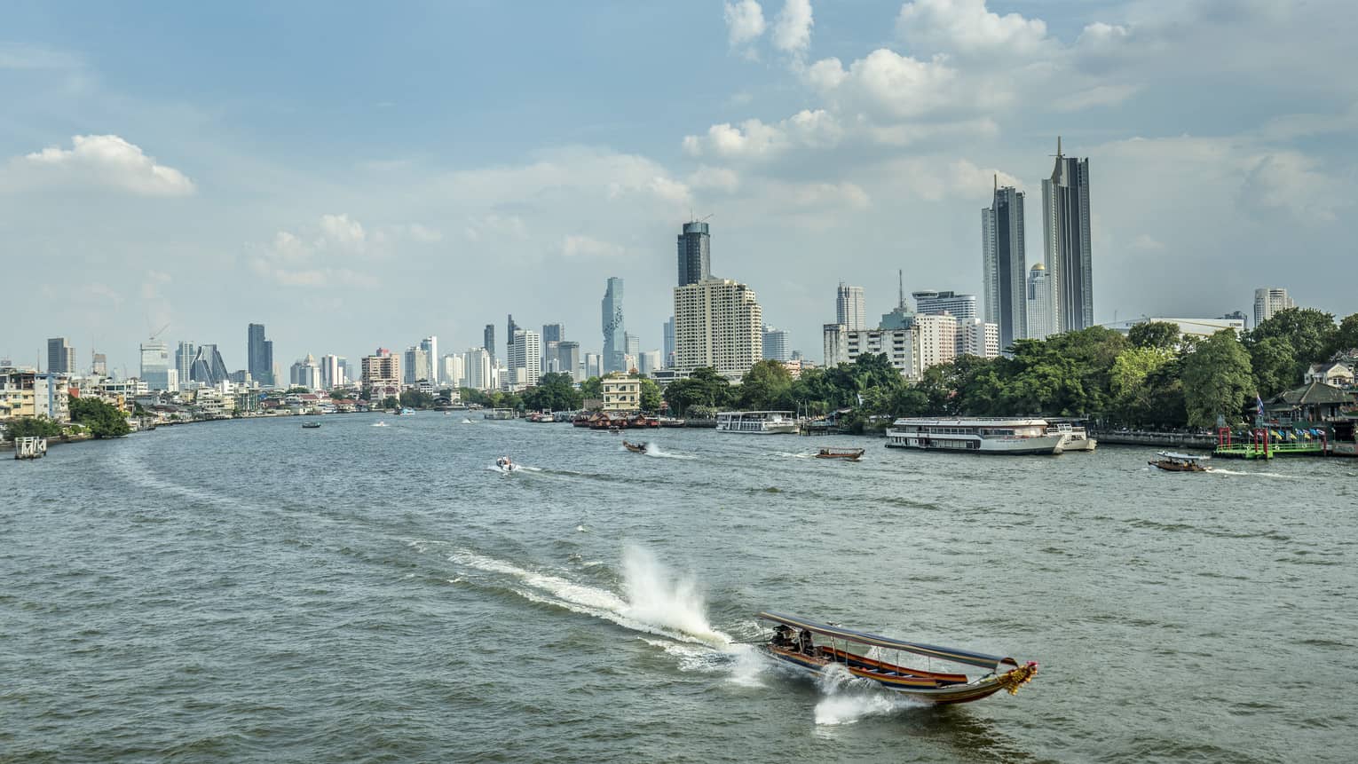 Longtail boat heading into foreground away from Bangkok skyline, leaving trail behind and surrounded by tall, splashing wake.
