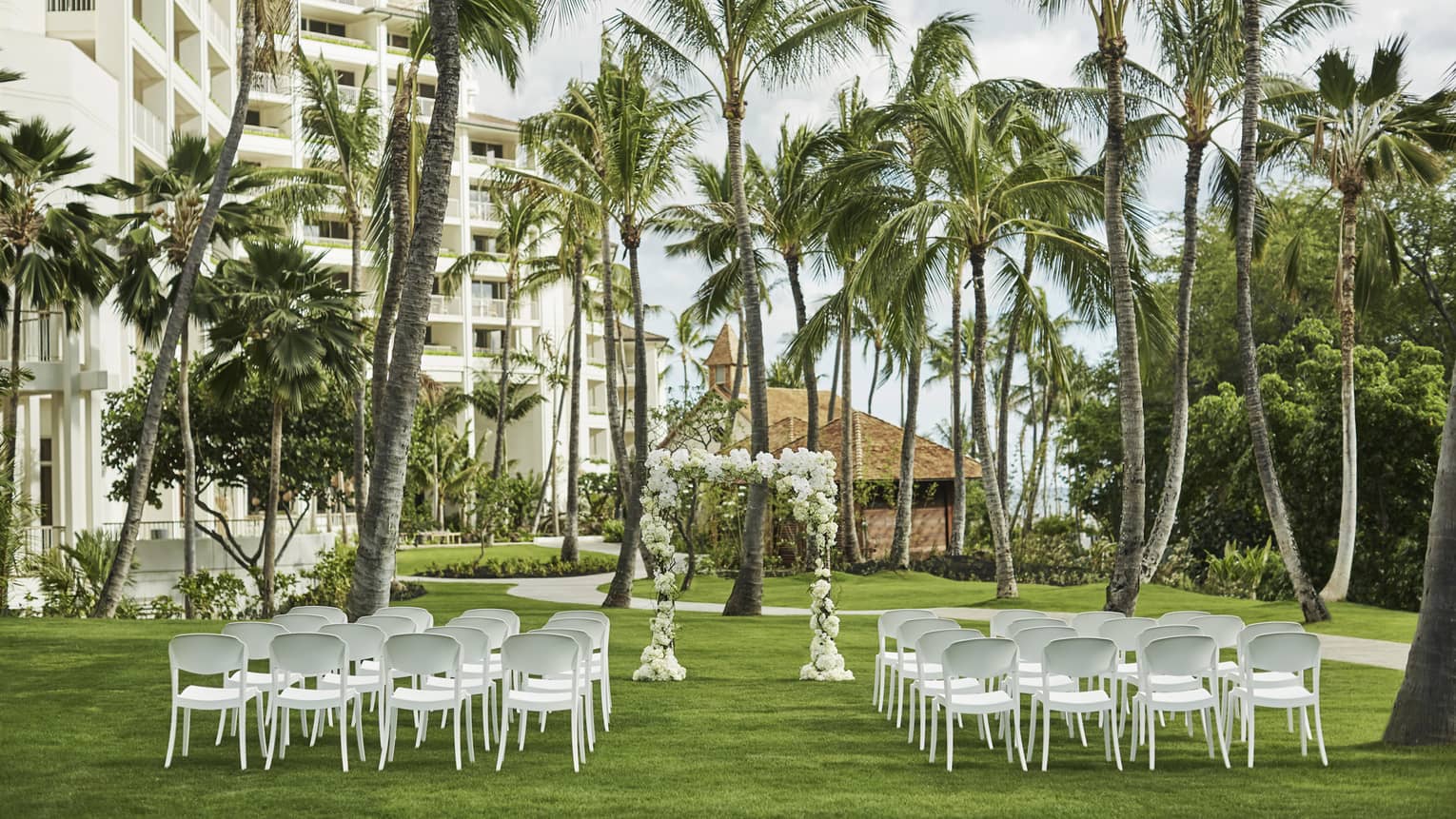 Outdoor wedding reception on event lawn, rows of white chairs face altar beneath palm trees