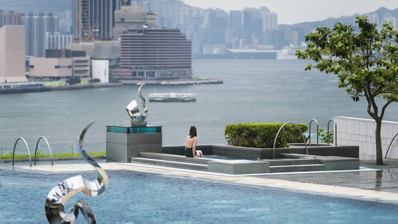 Woman in swimsuit sits on edge of swimming pool on patio near modern silver sculptures, waterfront