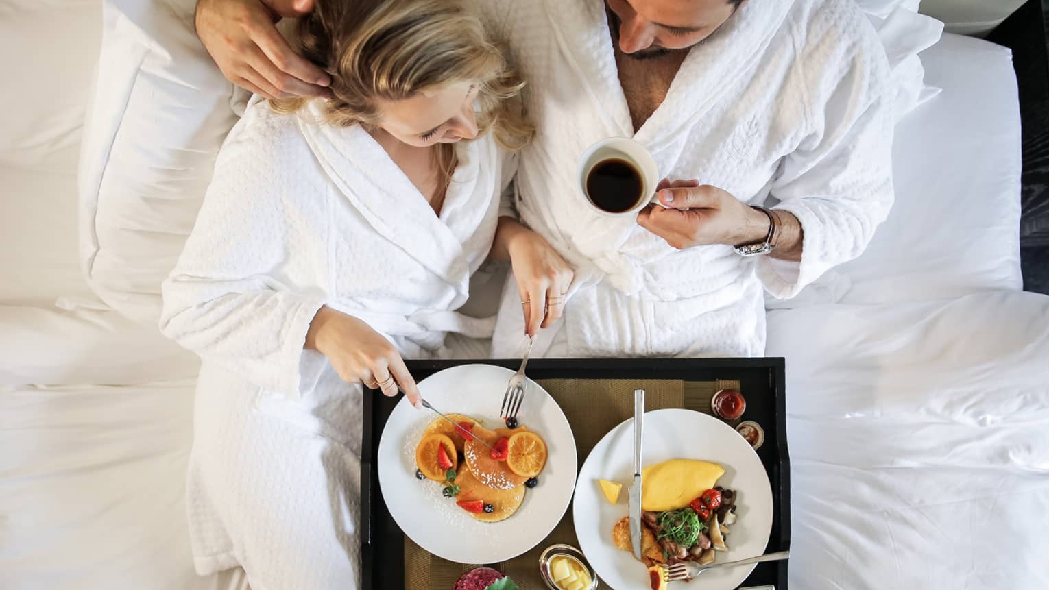 Couple wearing white robes cuddle in bed with breakfast tray