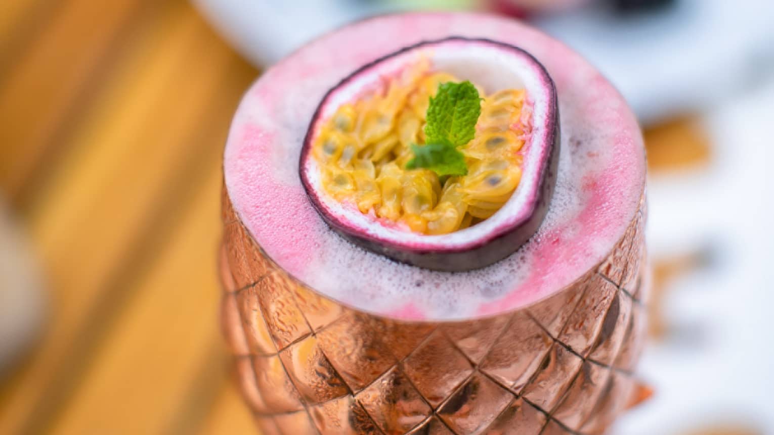 Pink cocktail in bronze pineapple-shaped goblet, garnished with passion fruit
