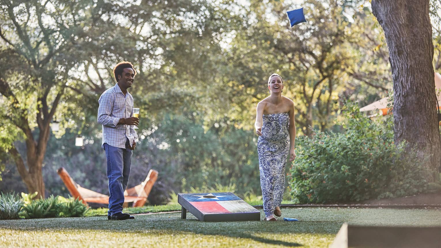 Laughing woman tosses bean bag on lawn, man holding beer watches