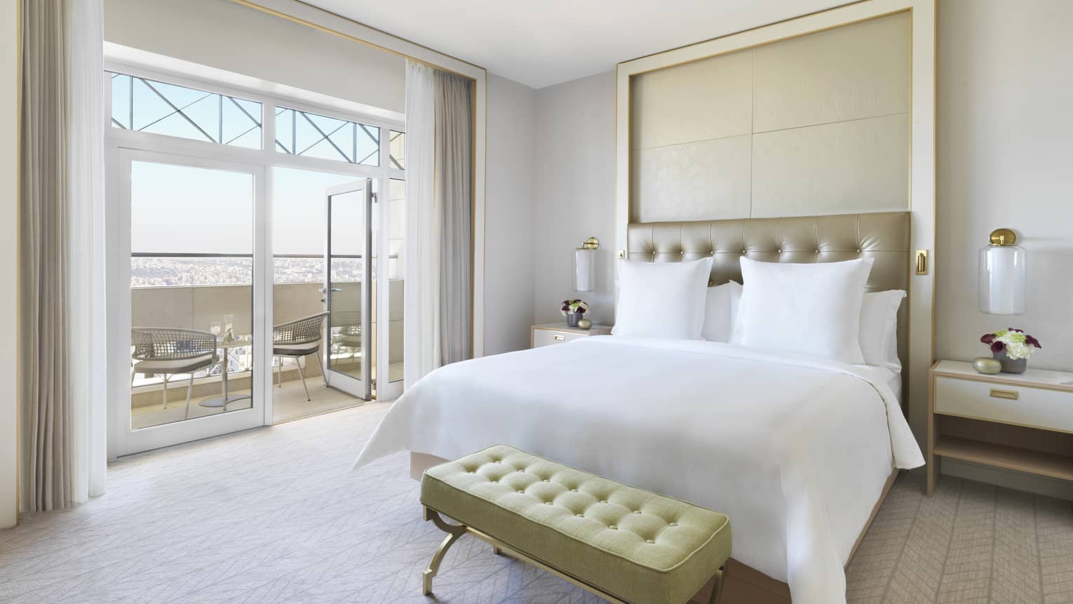 A plush white bed in the centre of the room with a yellow ottoman at the foot. The room opens up to a veranda overlooking the city. 