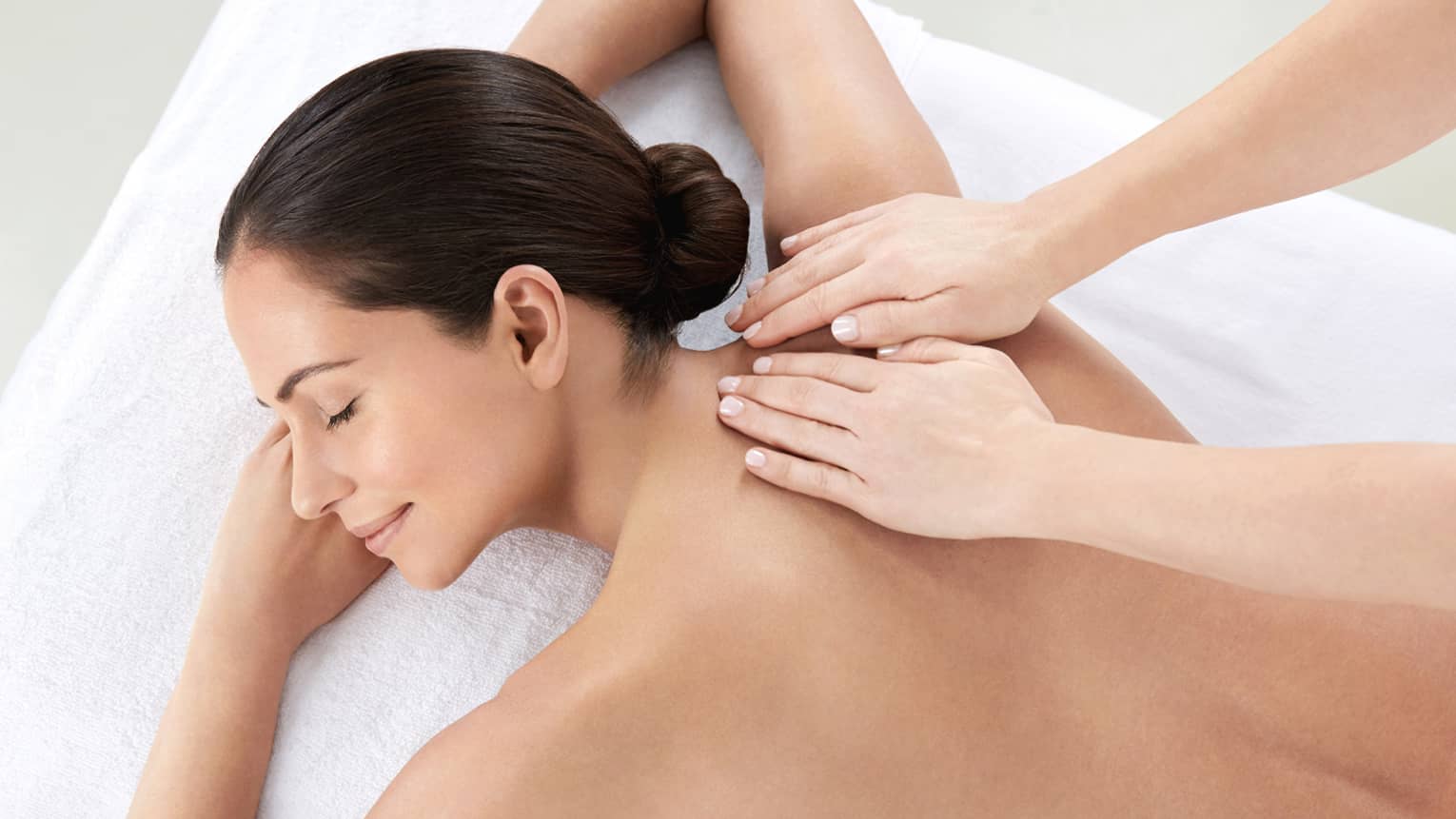 Woman smiles, lies on massage table as hands massage bare shoulders