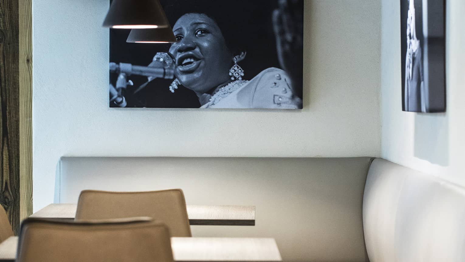 Two small tables with chairs and white banquette in modern bar with black fixtures, white walls and a hanging black-and-white portrait of Aretha Franklin 