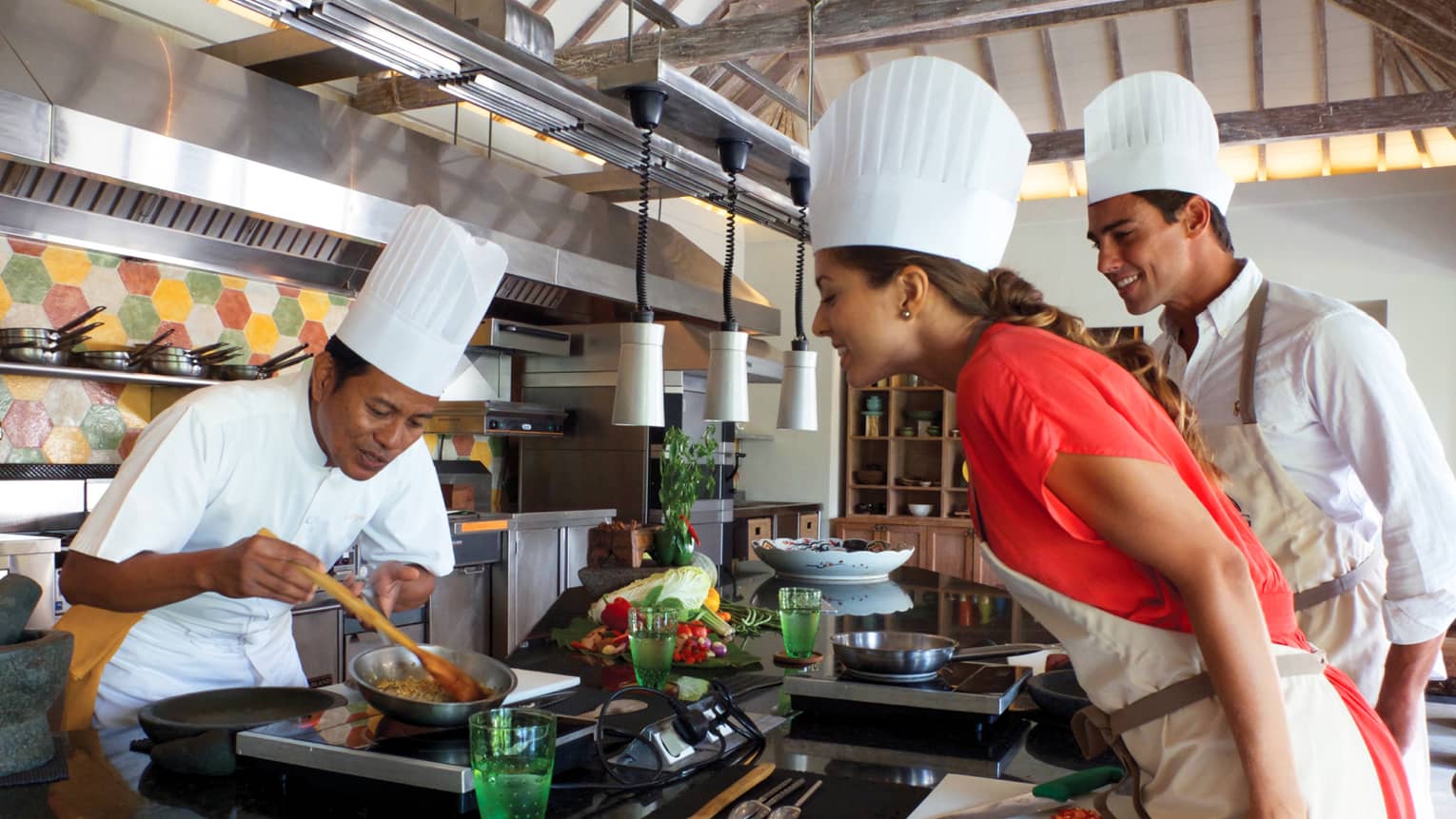 Chef in white hat, uniform stirs food in pan as woman and man in chef's hat, apron watch, smile