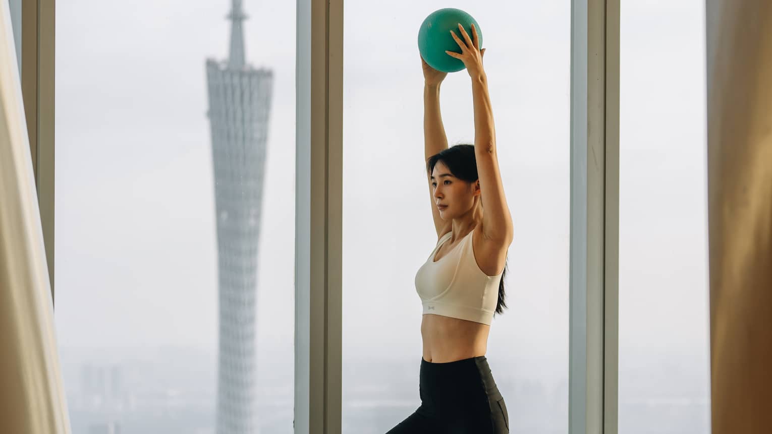 Guest holding a small exercise ball up above their head in front of floor-to-ceiling windows with a modern tower looming.