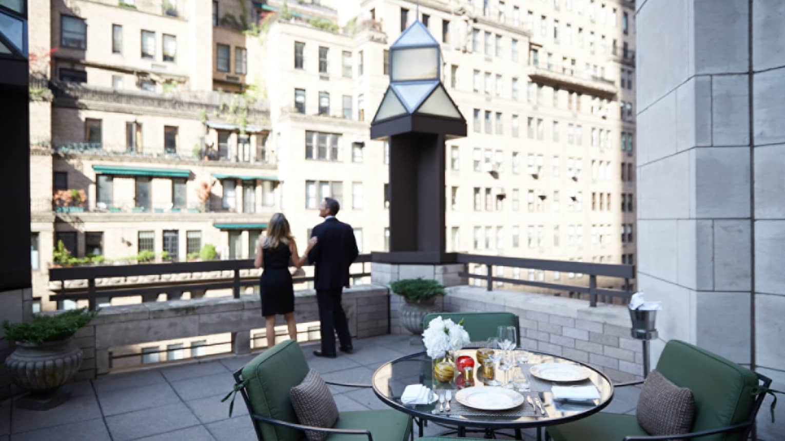 Man and woman in business suits look out from rooftop patio with table, plush chairs