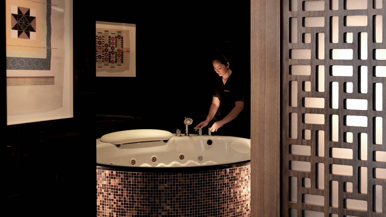 Woman holds taps of large round bathtub with tiles in dimly-lit spa room with wood room divider