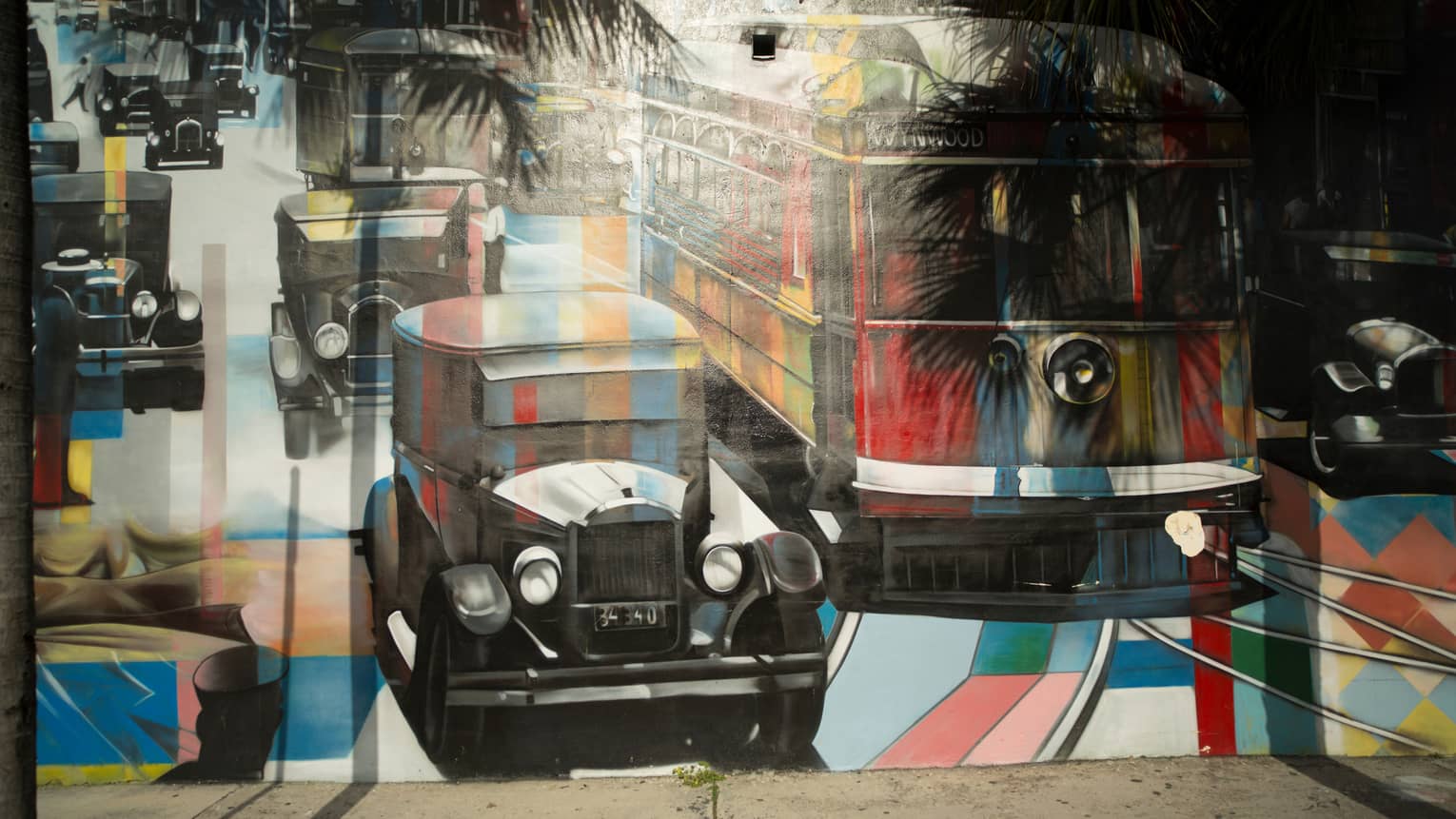 Palm trees cast shadows on a mural of vintage cars and a bus painted in a kaleiscopic pattern of colours and grey shading.