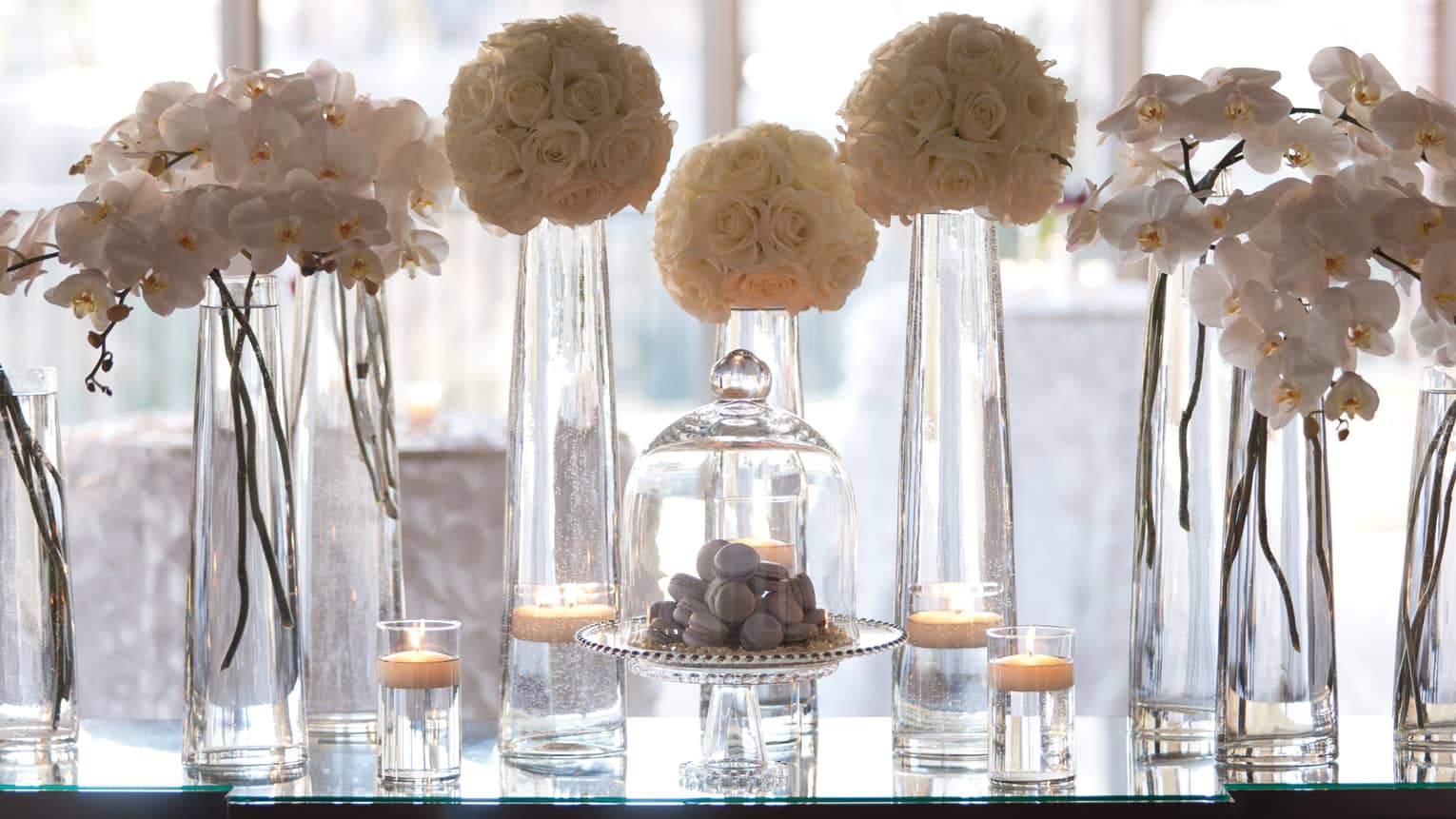 Flowers in tall glass vases, macarons in glass dessert display on table by window