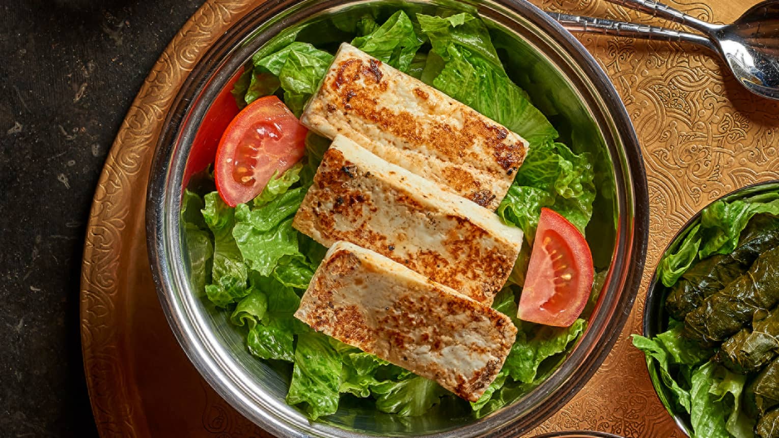 Three strips of fried Halloumi cheese on bed of lettuce in bowl, tomato wedge