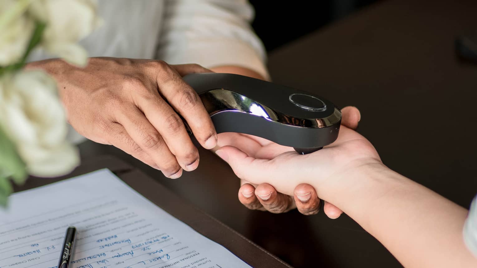 A person scans another's palm with a smooth, shiny, black electronic device beside a completed medical form on the table..