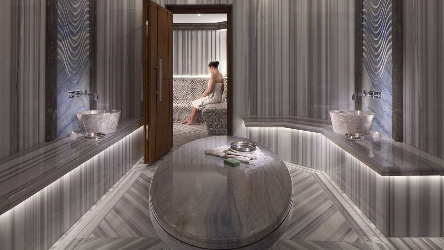 Elegant marble spa room with open wood door where woman wearing towel sits on bench 
