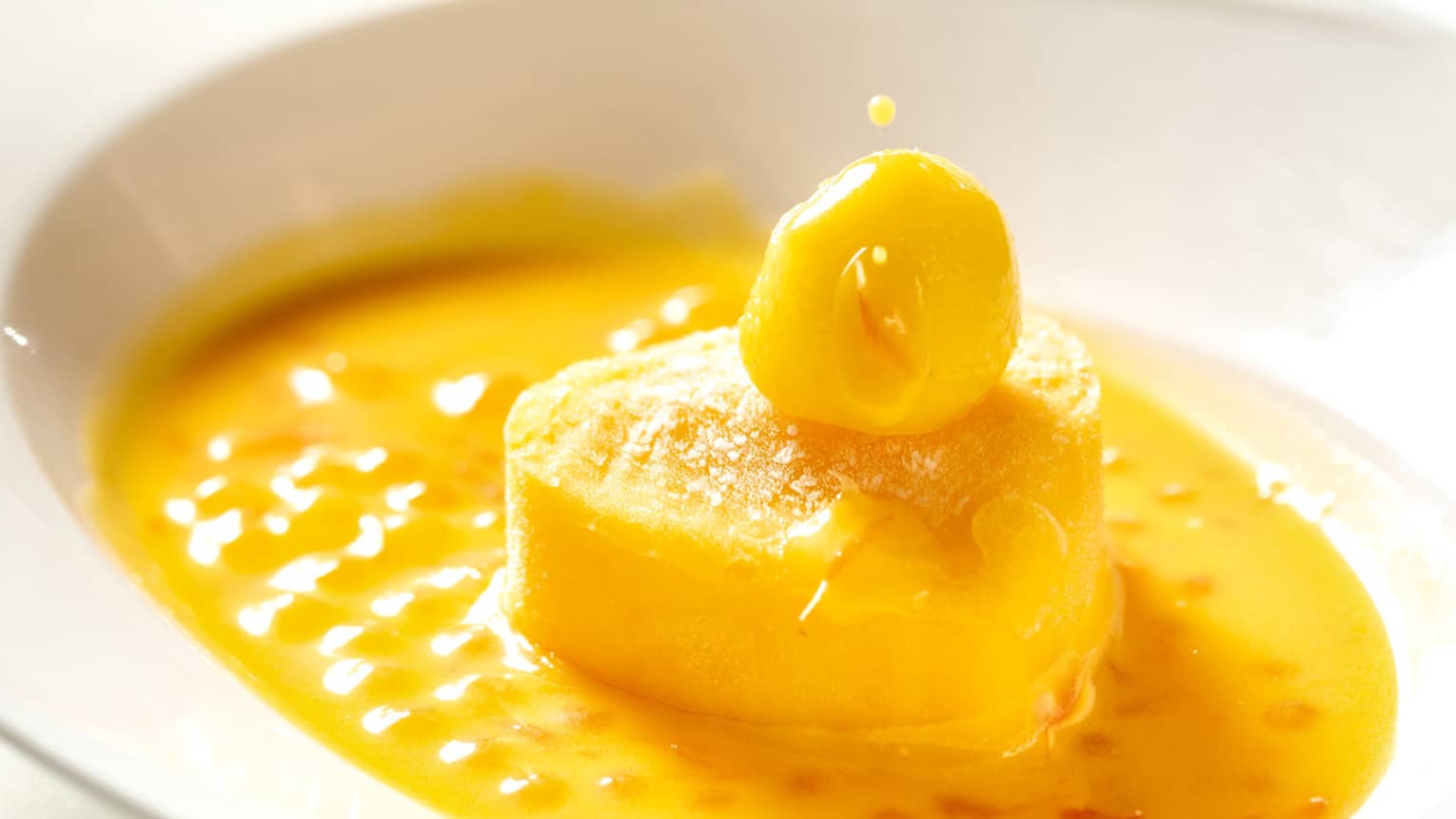 A white spoon drips yellow sauce onto a vibrant mango dessert served in a white dish