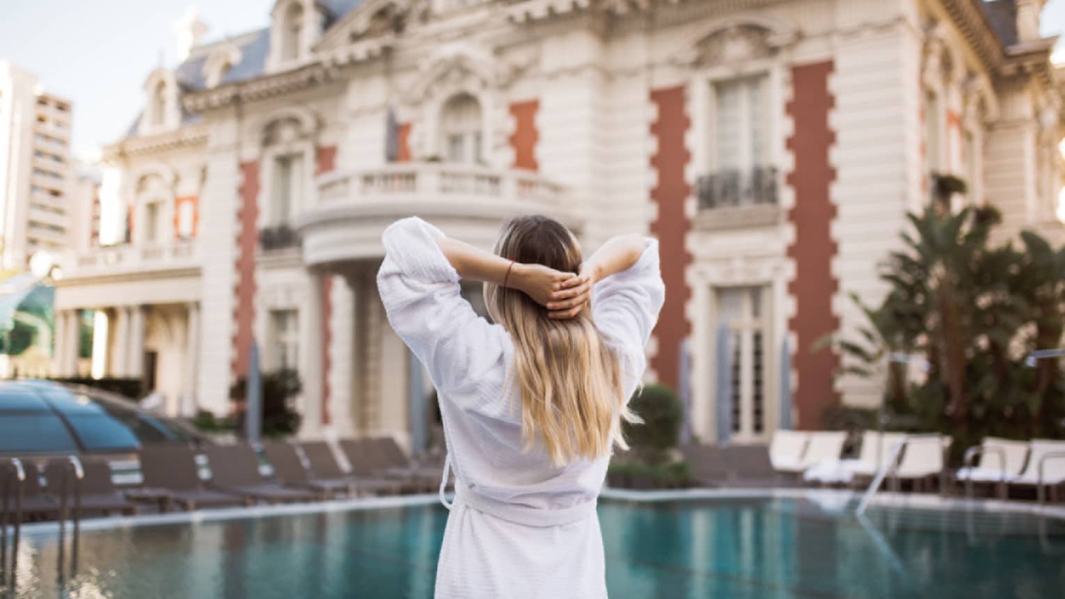 A woman in a white robe stretches by an outdoor pool, the hotel in the background