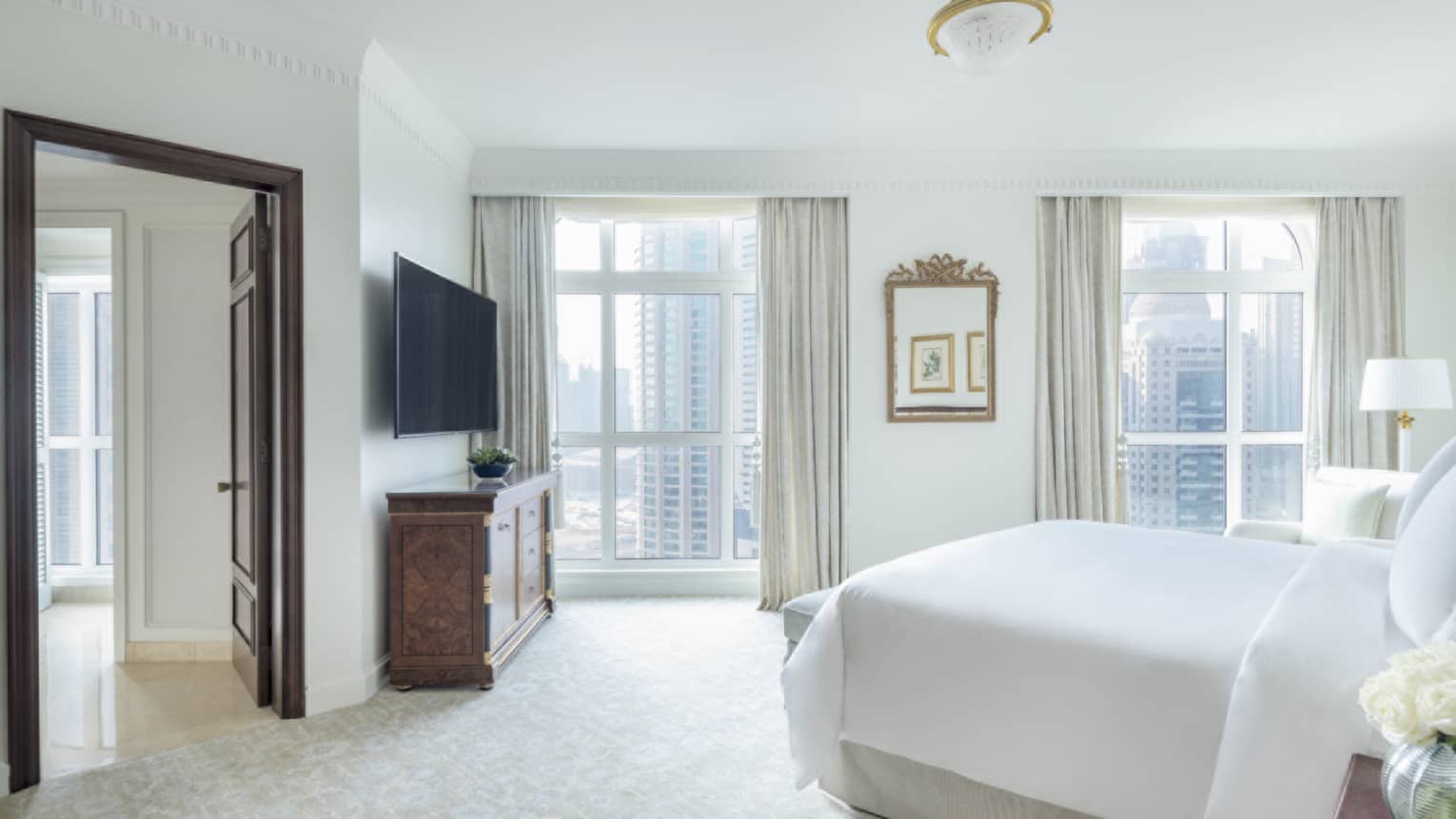 Light-filled bedroom with white bed, two floor-to-ceiling windows looking out to Doha city