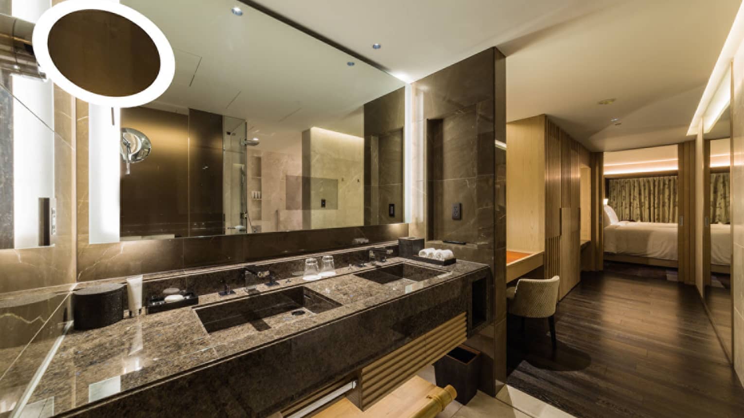 Bathroom with dark stone double vanity, illuminated mirror, open entrance to vanity and closet area as well as bedroom