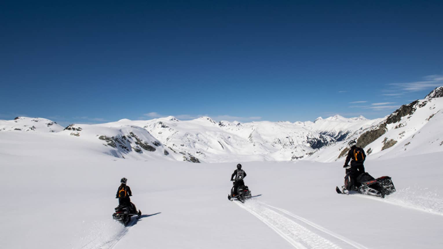 Three snowmobilers ride on pristine snow toward majestic snowy mountains and clear blue sky, leaving tracks in their wake.
