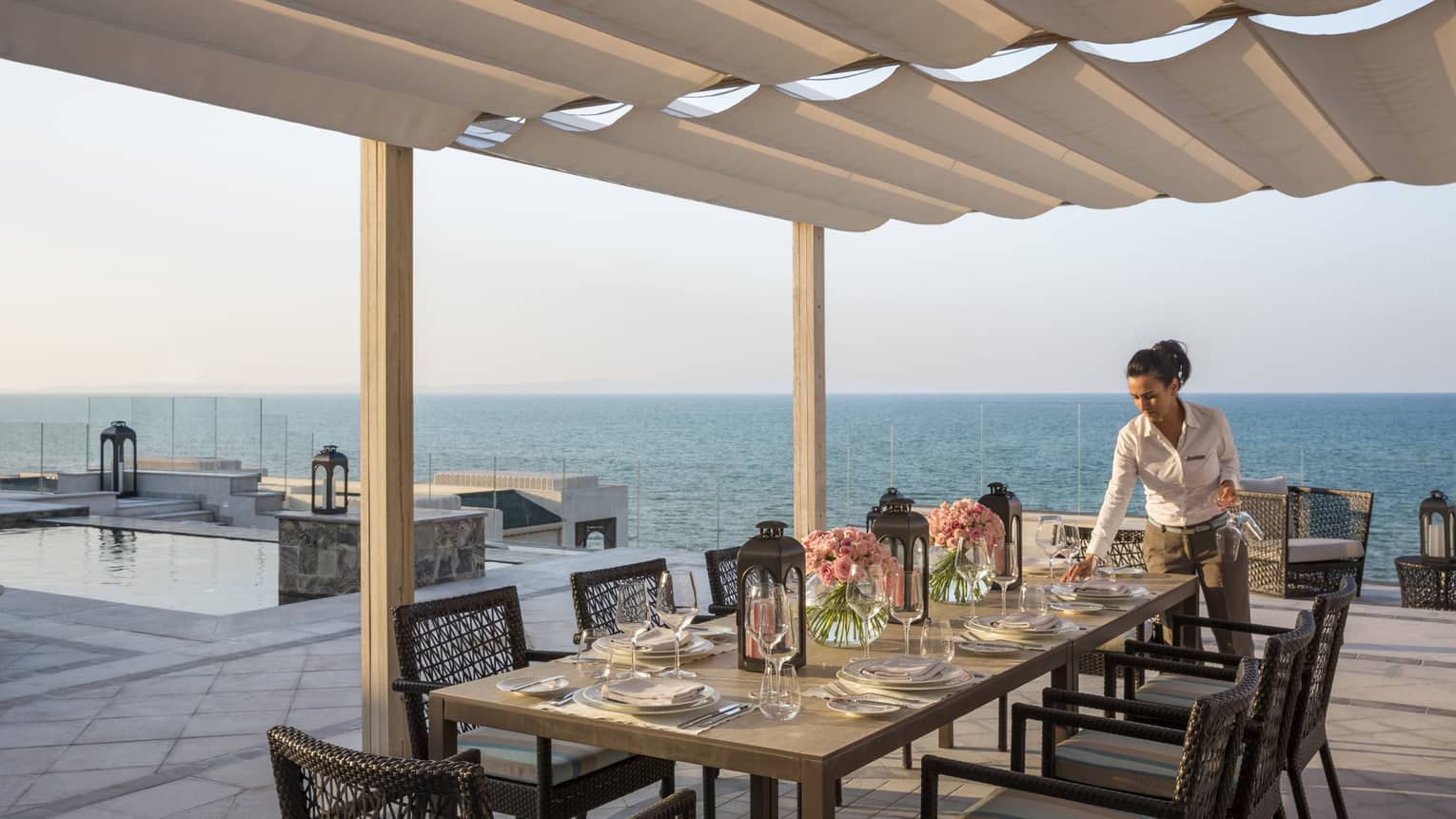 Woman sets table with flowers and lanterns on private rooftop patio overlooking pool and ocean views