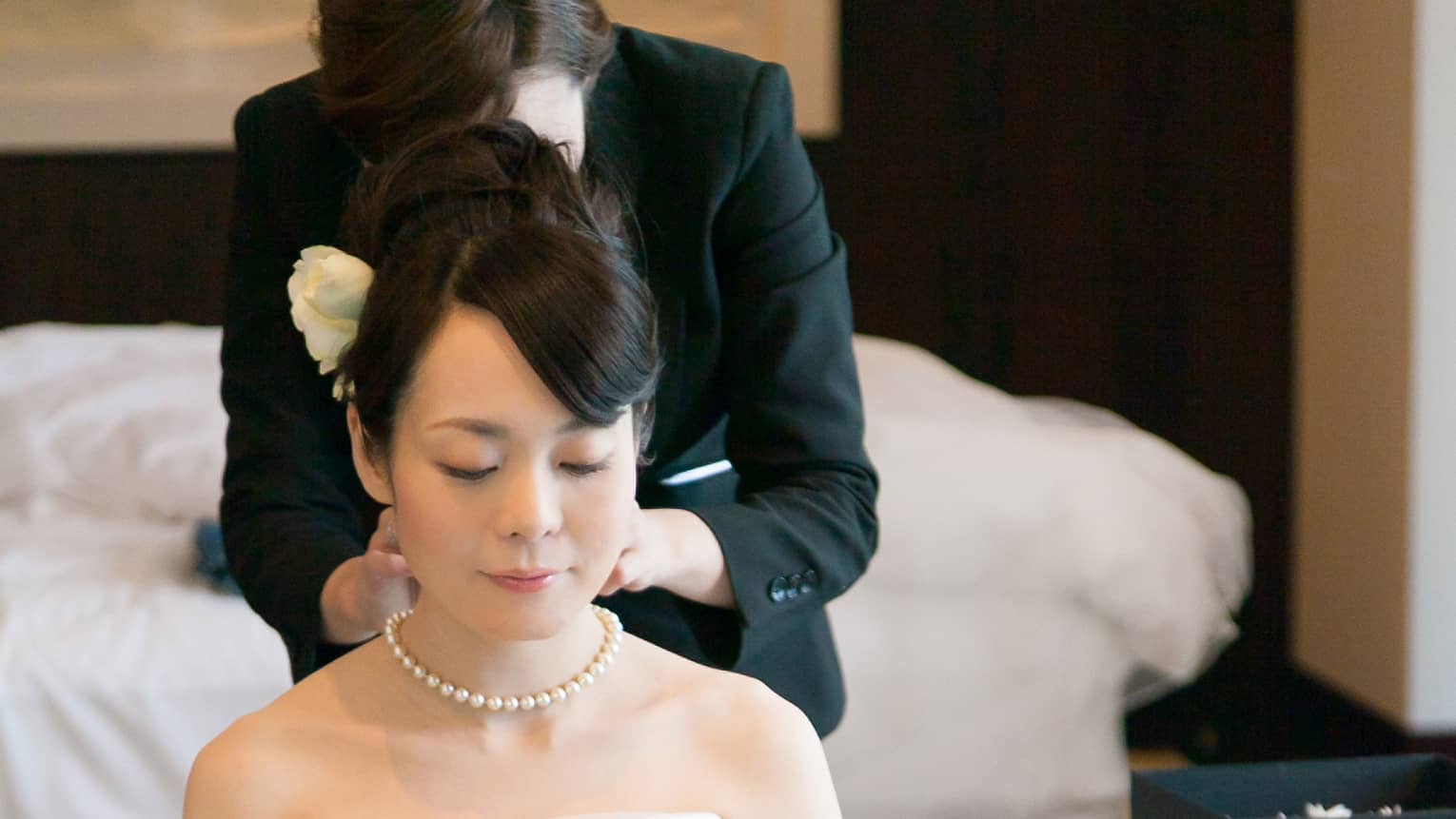 Hotel staff helps bride wearing wedding gown with her necklace in hotel room