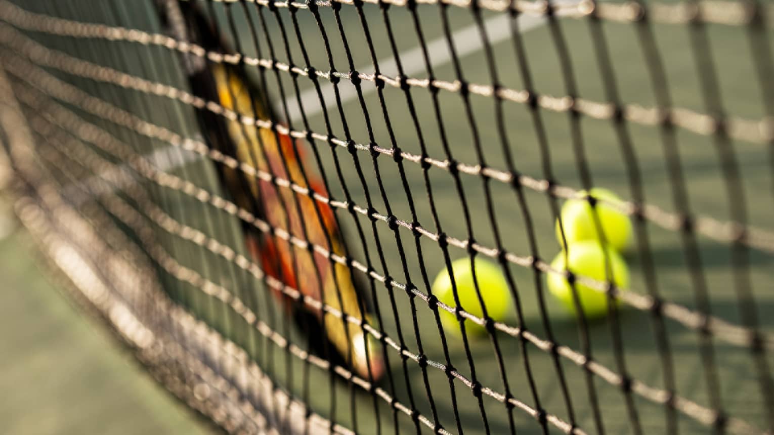 Close-up of tennis racket against net, three green tennis balls on ground