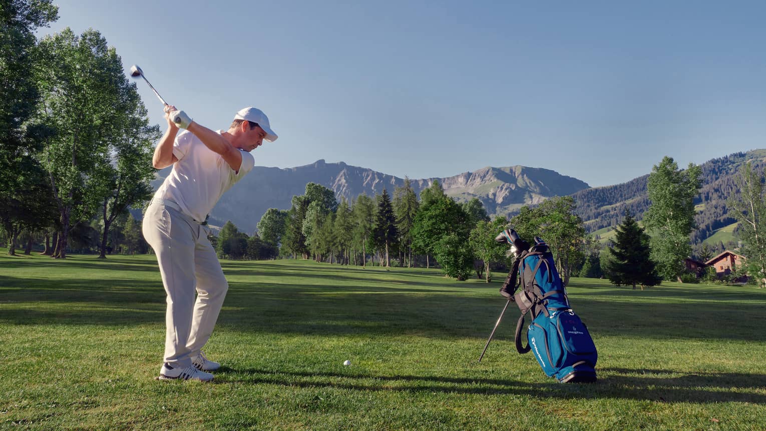 Man on mountainside golf course swings to hit golf ball beside blue golf bag