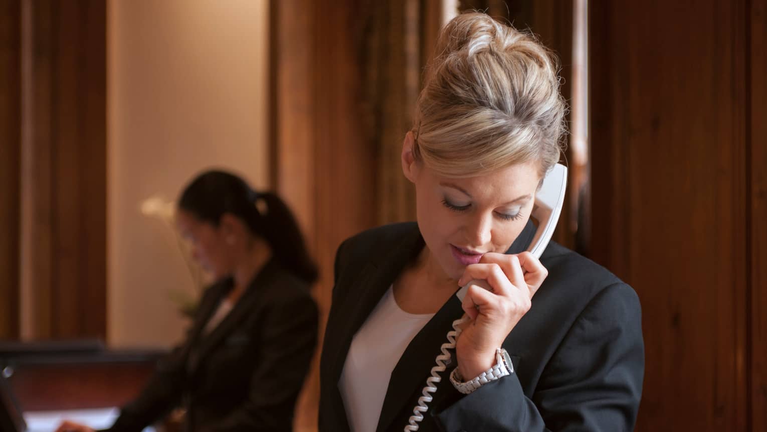 Close-up of woman in dark suit jacket holding telephone, cord, silhouette of hotel reception staff in background