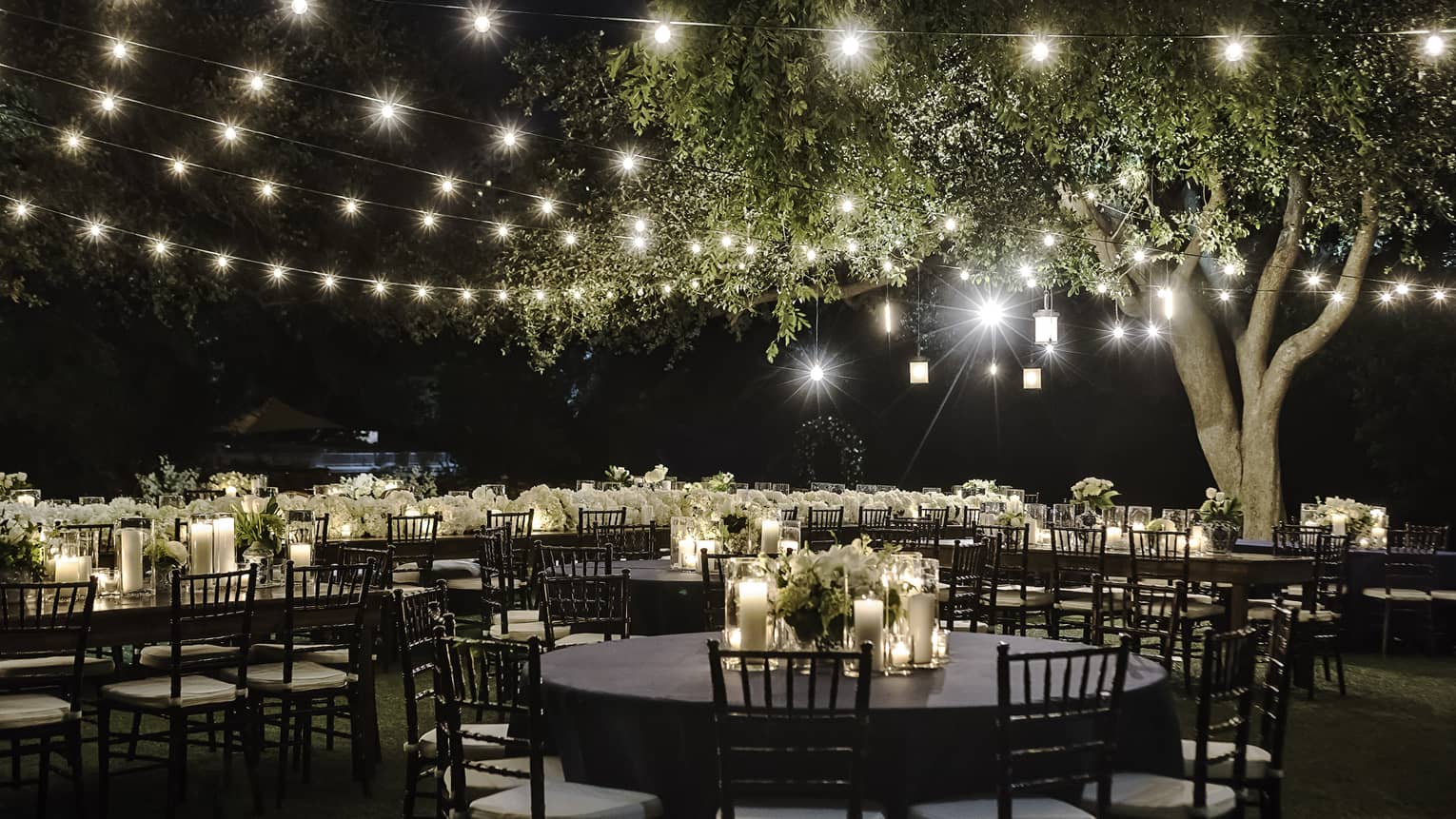 Lights strung across patio to large tree over round banquet tables with candles at night