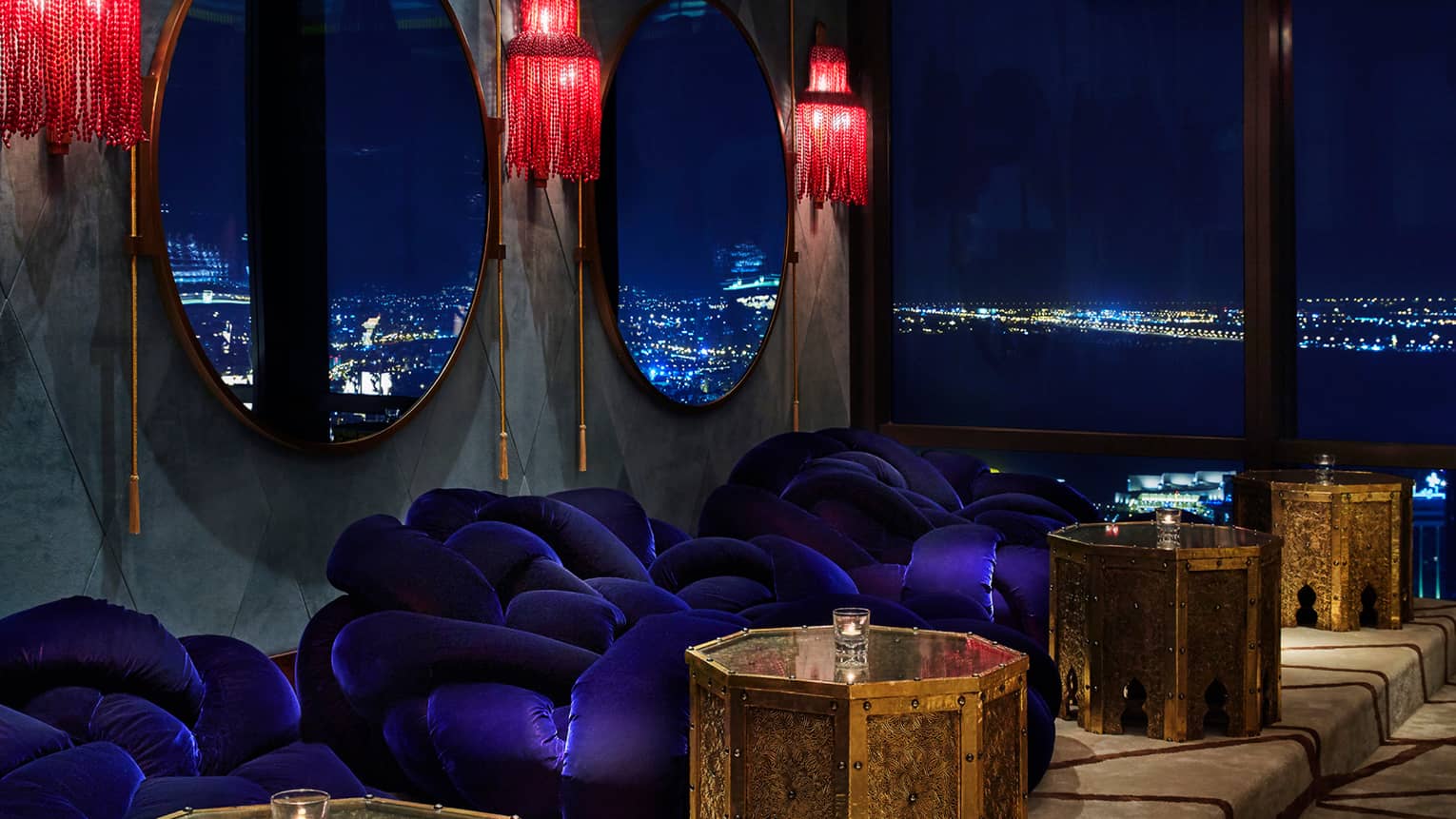 Close-up of blue velvet plush round chairs under red lamps, mirrors, gold tables, city lights