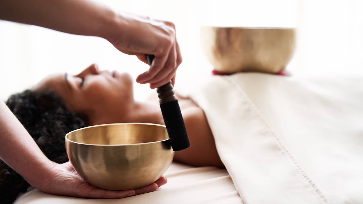 Woman with eyes closed lies on massage table in as vibrational sound therapy is performed with a Tibetan singing bowl